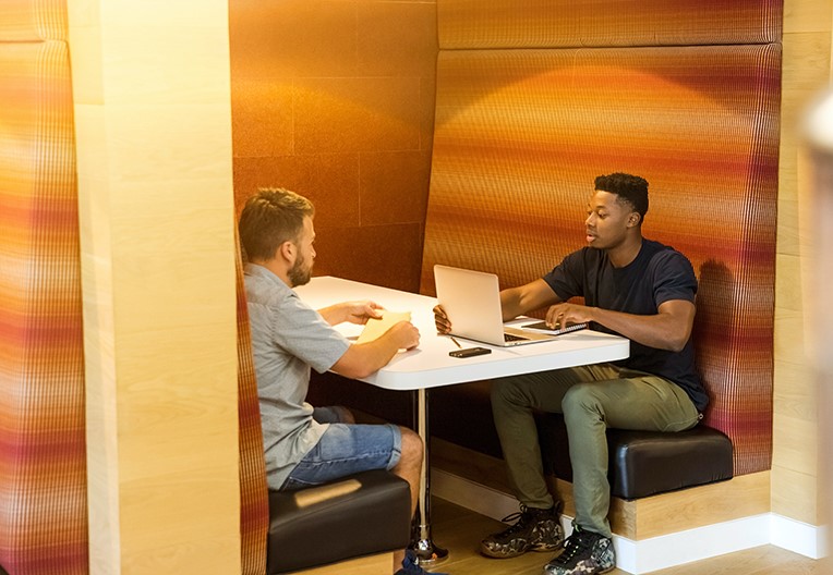 Two men sitting on a black leather bench holding grey laptop computer on top of white wooden table