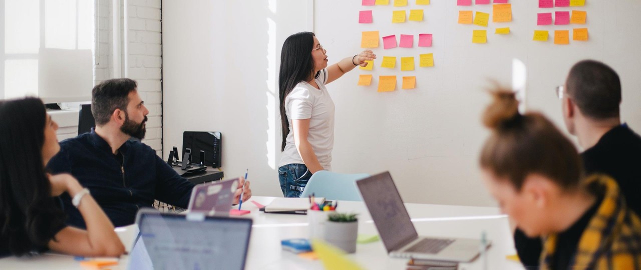 Woman placing sticky notes on the wall with people sitting around the table with their laptops having a meeting