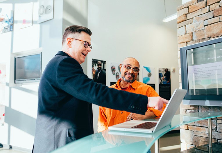 Two people are in discussion and smiling whilst one is pointing at a laptop