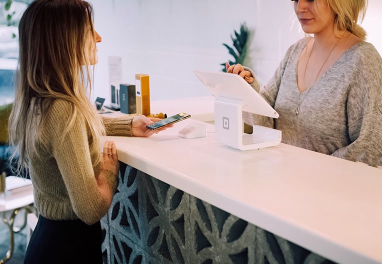 A woman holding her smartphone to make a payment with a card reader with another woman behind the counter