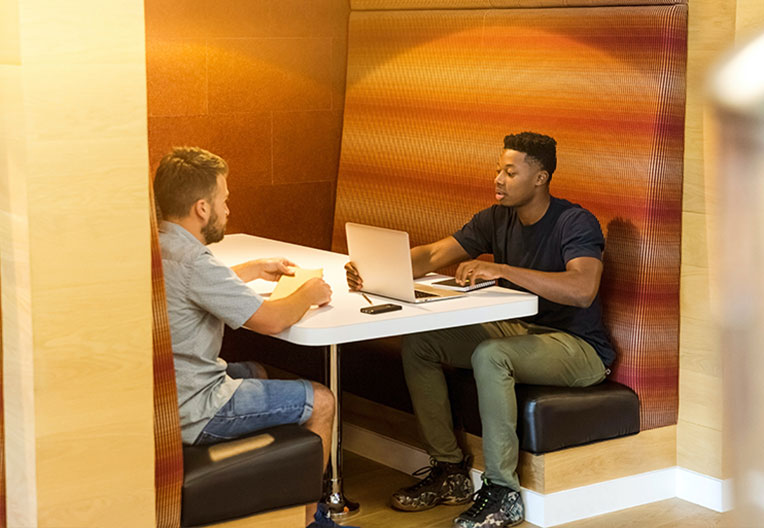 Two men sitting on a black leather bench holding grey laptop computer on top of white wooden table